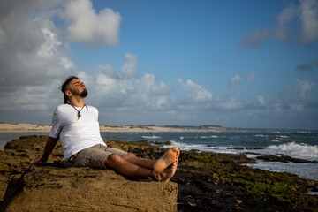 Young adult Caucasian man and hippie look relaxed on the rocks on the seashore during a sunrise.
