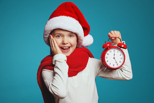 close up photo of excited pretty child in Santa hat with alarm clock isolated over blue background. Christmas countdown 