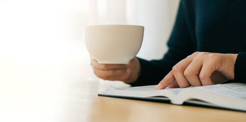 A young Christian woman sits at home reading and studying the Bible on Sundays with her morning coffee. beside the window sill