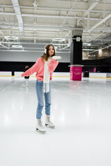 full length of happy woman in knitted sweater, ear muffs and winter outfit skating while sending air kiss on ice rink.