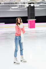 full length of happy woman in knitted sweater, ear muffs and winter outfit skating with paper cup on ice rink.