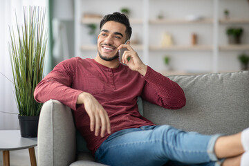 Fototapeta premium Happy middle-eastern guy reclining on couch, talking on phone
