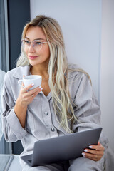 nice caucasian female in casual domestic clothing using computer laptop and sitting on window sill alone at home, drinking coffee. Freelance working, free time and leisure, online browsing internet