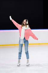 full length of happy woman in ear muffs and winter outfit skating on ice rink.