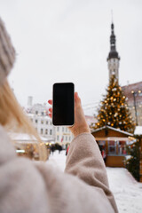A girl holds a smartphone in her hands and takes pictures of a decorated Christmas tree on the street to share photos on social networks. Festive mood and atmospheres. Christmas tree