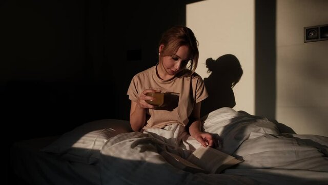 beautiful woman sitting in bed and reading a book in her pajamas with juice in her hand on sunny morning, morning sunlight reading routine