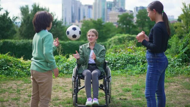 Smiling cheerful beautiful disabled girl in wheelchair playing ball with friends in summer park with blurred urban city at background. Portrait of happy Caucasian child enjoying leisure outdoors