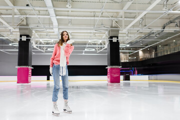 full length of happy young woman in winter outfit skating and sending air kiss on ice rink.