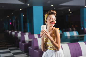 cheerful pin up woman in dress holding tasty milkshake in cafe.
