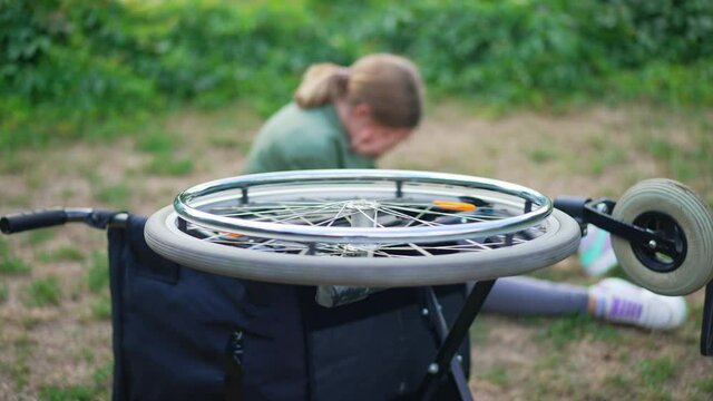 Wheel of rolled over wheelchair spinning with blurred disabled girl crying at background. Frustrated helpless hopeless Caucasian teenager in summer park outdoors alone. Depression concept