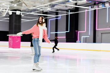 full length of cheerful young woman in winter outfit skating with outstretched hands on ice rink.