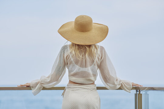Stunning blonde woman in summer beach outfit relaxing outdoors against sea background. Back view of fashionable romantic young adult lady wearing a trendy vintage straw hat, white blouse, and skirt