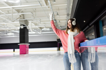 happy woman in white gloves, ear muffs and pink sweater waving hand on ice rink.