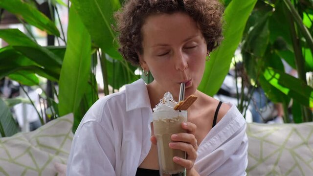 Happy eco-friendly young adult woman drinking ice latte with cream and cookies on the top using metal straw 