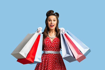 Excited young pinup woman in red polka dot dress holding lots of shopping bags on blue studio...