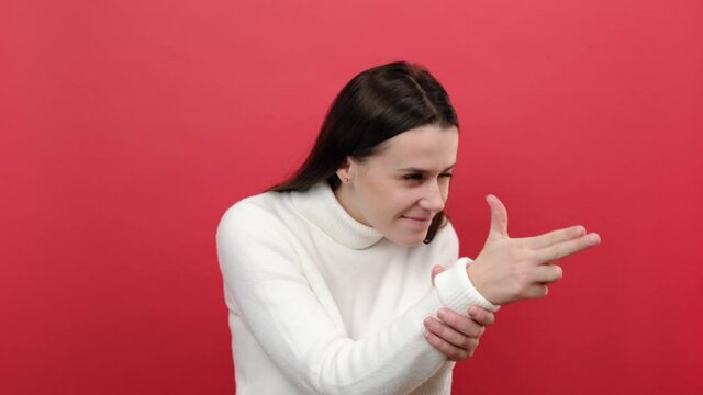 Portrait of young female 20s old years points finger gun, threatening to kill, shoots with weapon gesture right in target, dressed in white knitted sweater, posing isolated over red color background
