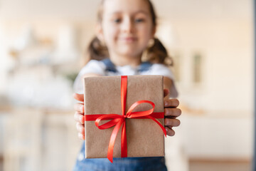 Small cute daughter girl holding focused present gift box with red ribbon, giving receiving...