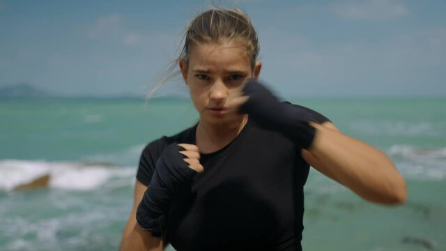 Sporty young boxer girl in black top has boxing training on the ocean background in sunny morning