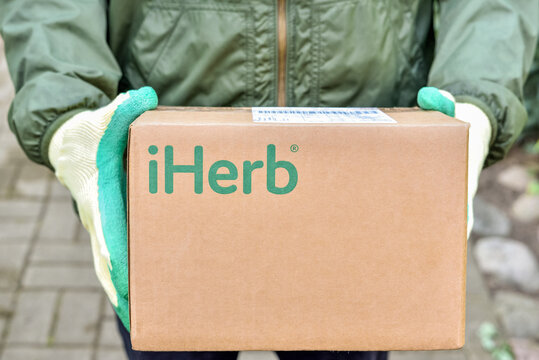 Logotype iHerb on cardboard box in courier hands. iHerb is an herbal products company, provides and delivers health and wellness products to people around the world: Riga, Latvia - September 10, 2021