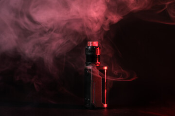 Electronic vaping device mod, atomizer stands in vape smoke and vapor illuminated by red light.