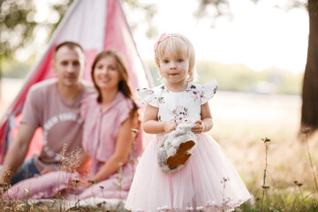 Mom, dad and little daughter are sitting next to wigwam decoration in the park. Family spending time outdoor in summer, having fun together. Girl are dressed in pink dress. © Andriy Medvediuk