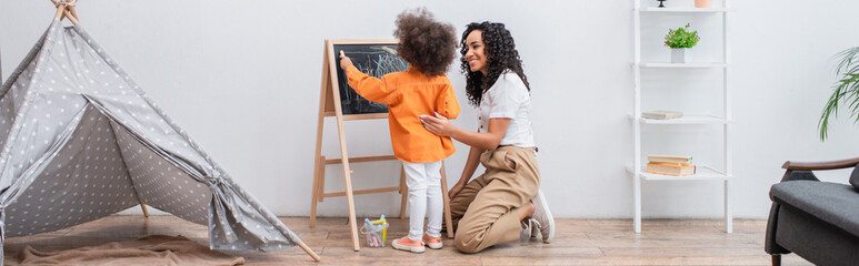 Smiling mother looking at african american daughter drawing on chalkboard near tent at home, banner.