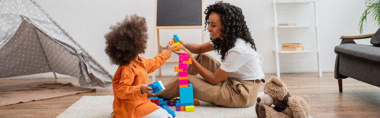 Smiling african american mother playing colorful building blocks near teepee at home, banner. © LIGHTFIELD STUDIOS