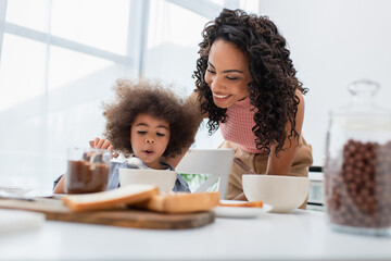Smiling african american parent standing near daughter eating breakfast near cereal and chocolate paste.