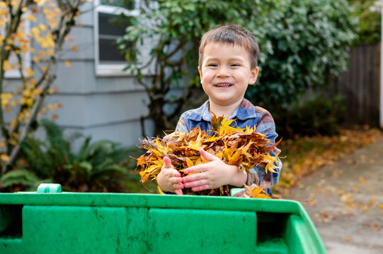 Young boy smiling with pile of leaves in arms and bin in front