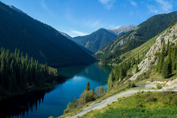 In a beautiful alpine gorge there is a large lake Kolsai, the lake is surrounded by mountain ranges with peaks, a spruce forest grows on the slopes, a road in the foreground, summer, sunny