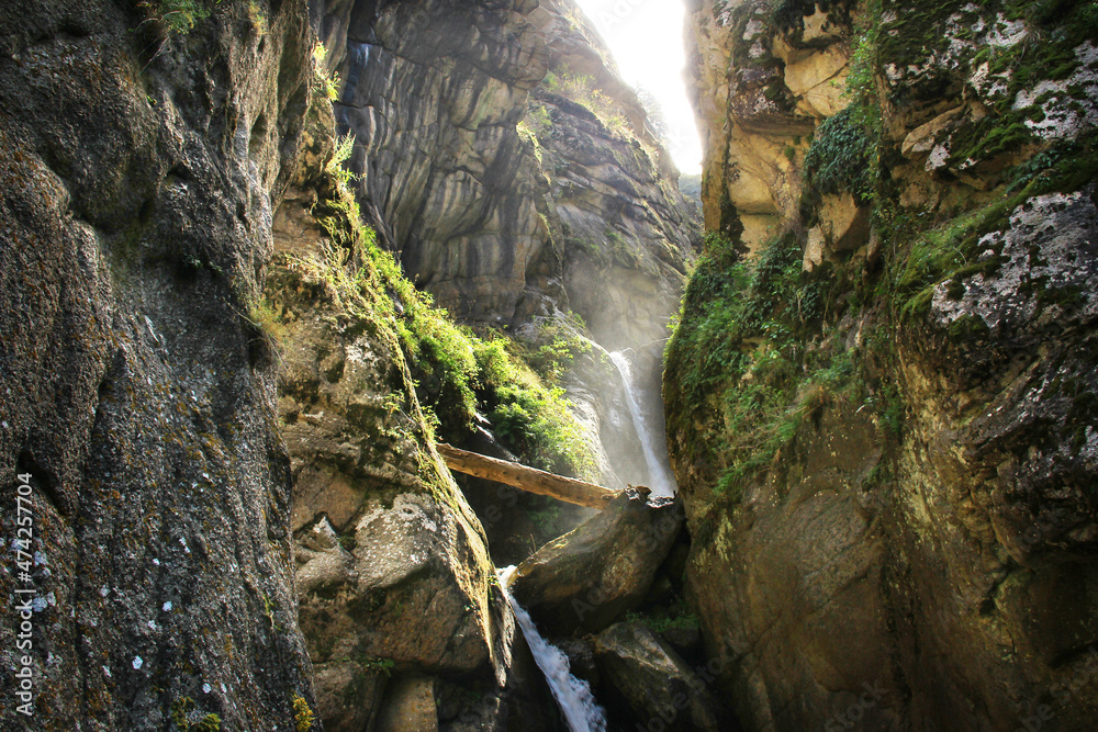 Wall mural Waterfall in the mountains between the rocks. Sunny day in the mountains, rocks, gray stones covered with moss, a waterfall flows in a stream between huge stones, grass grows on the stones