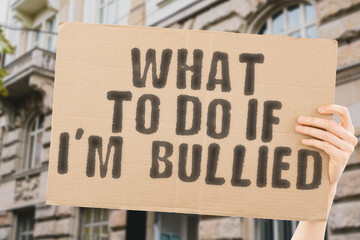 The phrase " What to do if I’m bullied " on a banner in men's hand with blurred background. Anxiety. Adolescence. Society. Abused. Anger. Friends. Troubled. Stressful. Discrimination