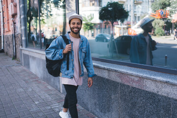 Portrait of trendy dressed Latino student walking at city streets and smiling at camera during free...