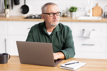 Elderly man working at laptop computer sitting at the table  in the home kitchen