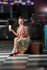 happy pin up woman holding milkshake while sitting on retro suitcase in cafe.