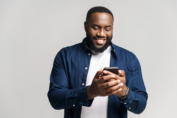 Optimistic African-American man wearing casual shirt texting on the smartphone, texting, smiling black guy enjoying using new mobile app isolated on grey