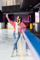 positive young woman with paper cup taking selfie on ice rink.