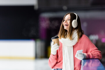 excited woman in ear muffs holding paper cup and smartphone on ice rink.