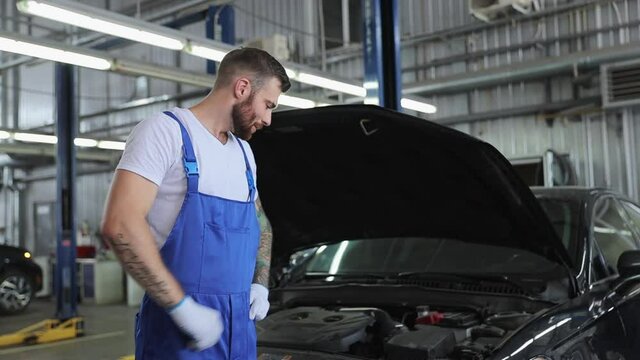 Pensive professional technician young brunet car mechanic man 20s wears denim overalls white t-shirt gloves working in vehicle repair shop with open hood on light modern workshop background indoors