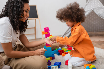 Side view of smiling african american woman playing building blocks near daughter at home.