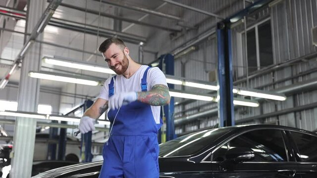 Vivid overjoyed young brunet car mechanic man 20s wears denim overalls white t-shirt gloves headphones working in vehicle repair shop inspecting hold wrench dancing modern workshop background indoors