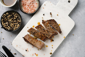 A piece of cooked beef meat steak on white marble board cut in slices, white and black peer, coarse sea salt