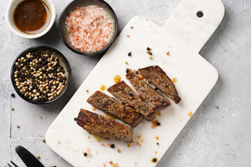 A piece of cooked beef meat steak on white marble board cut in slices, white and black peer, coarse sea salt