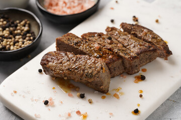 A piece of cooked beef meat steak on white marble board cut in slices, white and black peer, coarse sea salt