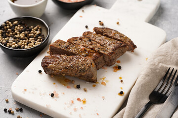 A piece of cooked beef meat steak on white marble board cut in slices, white and black peer, coarse sea salt