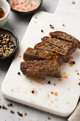 A piece of cooked beef meat steak on white marble board cut in slices, white and black peer, coarse sea salt