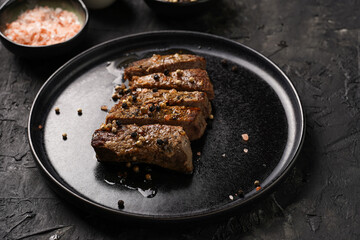 A piece of cooked beef meat steak on black plate cut in slices, white and black peer, coarse sea salt
