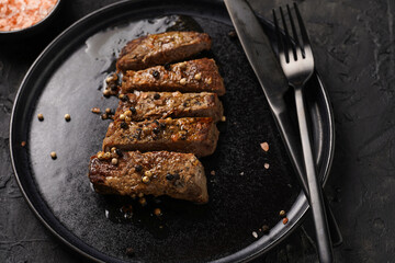 A piece of cooked beef meat steak on black plate cut in slices, white and black peer, coarse sea salt