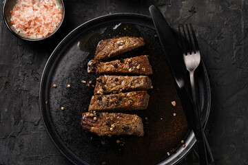 A piece of cooked beef meat steak on black plate cut in slices, white and black peer, coarse sea salt