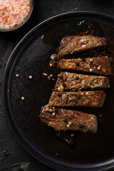 A piece of cooked beef meat steak on black plate cut in slices, white and black peer, coarse sea salt